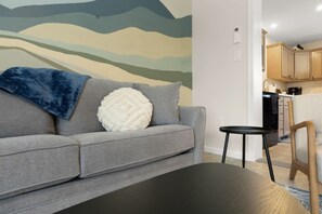 Uma sala de estar com um sofá cinza, uma almofada branca texturizada, um cobertor azul, uma mesa de centro preta e uma parede com um mural de padrão de onda.