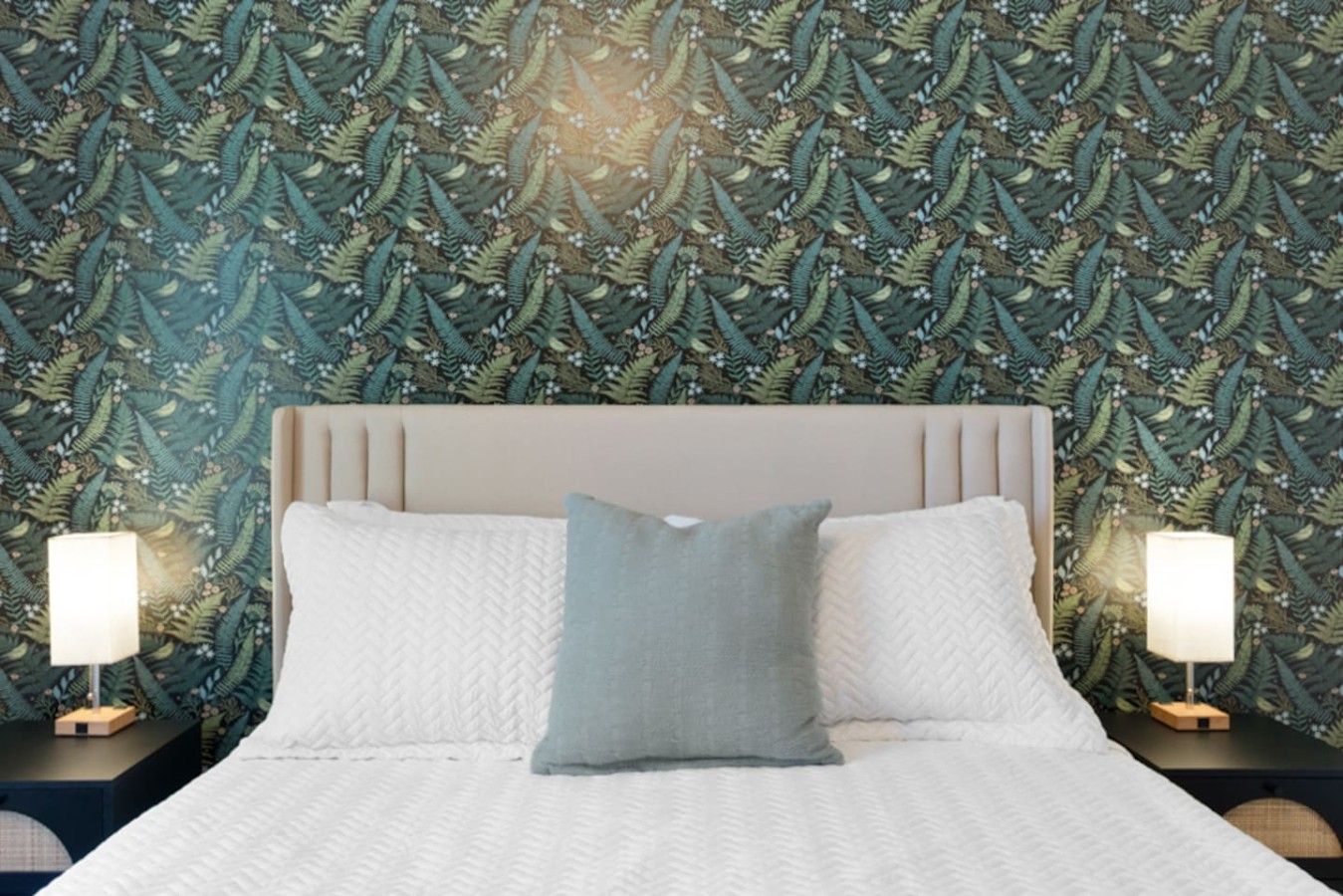 A bed with a white quilt and two pillows, flanked by nightstands with lamps, against a wall with a leafy pattern.