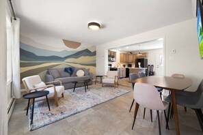 Uma sala de estar moderna com sofá, cadeiras, mesa de centro e mesa de jantar. Cozinha ao fundo, com armários e eletrodomésticos.