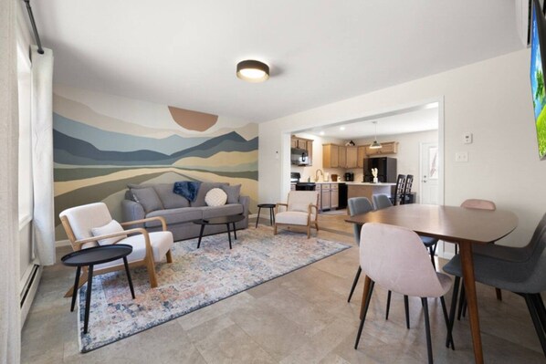 A modern living room with a sofa, chairs, a coffee table, and a dining table. There is a kitchen in the background with cabinets and appliances.