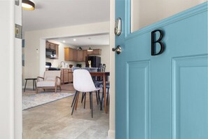 Quarto com porta azul marcada com a letra 'B', área de jantar com cadeiras e mesa, e cozinha ao fundo.