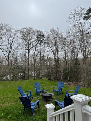 A grassy outdoor area with a white railing, surrounded by trees and blue chairs arranged around a central table. - Maine Forest Hideaway (Ellsworth)