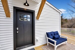 A white building with a black door, a small window, and a blue cushioned bench on a wooden deck.