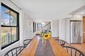A long dining table with chairs, a vase with flowers, and a kitchen area with a refrigerator.