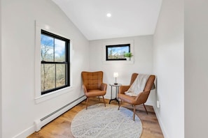 A room with a large window, two leather chairs, a small table with a lamp, and a round rug.