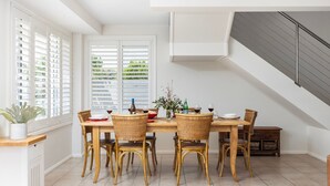 A dining area with a wooden table and wicker chairs, large windows with blinds, and a staircase with a grey railing. - Swell – walk to Fingal Bay Beach, swimming pool, outdoor BBQ entertaining (Fingal Bay)