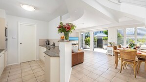 A spacious kitchen with a central island, a dining area with wicker furniture, and a view of the outdoors through large windows. - Swell – walk to Fingal Bay Beach, swimming pool, outdoor BBQ entertaining (Fingal Bay)