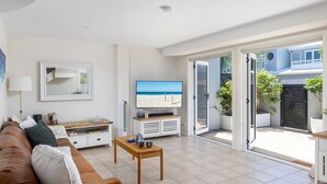 A modern living room with a flat-screen TV, a sofa, a coffee table, and a sliding glass door leading to a balcony with plants. - Swell – walk to Fingal Bay Beach, swimming pool, outdoor BBQ entertaining (Fingal Bay)