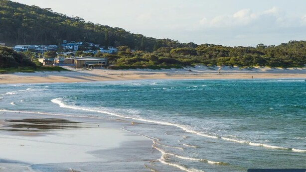A beach with clear blue waters, sandy shore, and houses in the background. - Swell – walk to Fingal Bay Beach, swimming pool, outdoor BBQ entertaining (Fingal Bay)