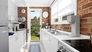 Een compacte keuken met witte kastjes, een magnetron, waterkoker en koffieapparaat. Er is een bakstenen muur en een raam met uitzicht op groen.