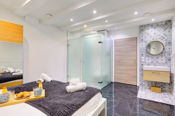 Una habitación de hotel moderna con una cama, cabina de ducha de vidrio, baño con lavamanos y una bandeja de desayuno sobre la cama.