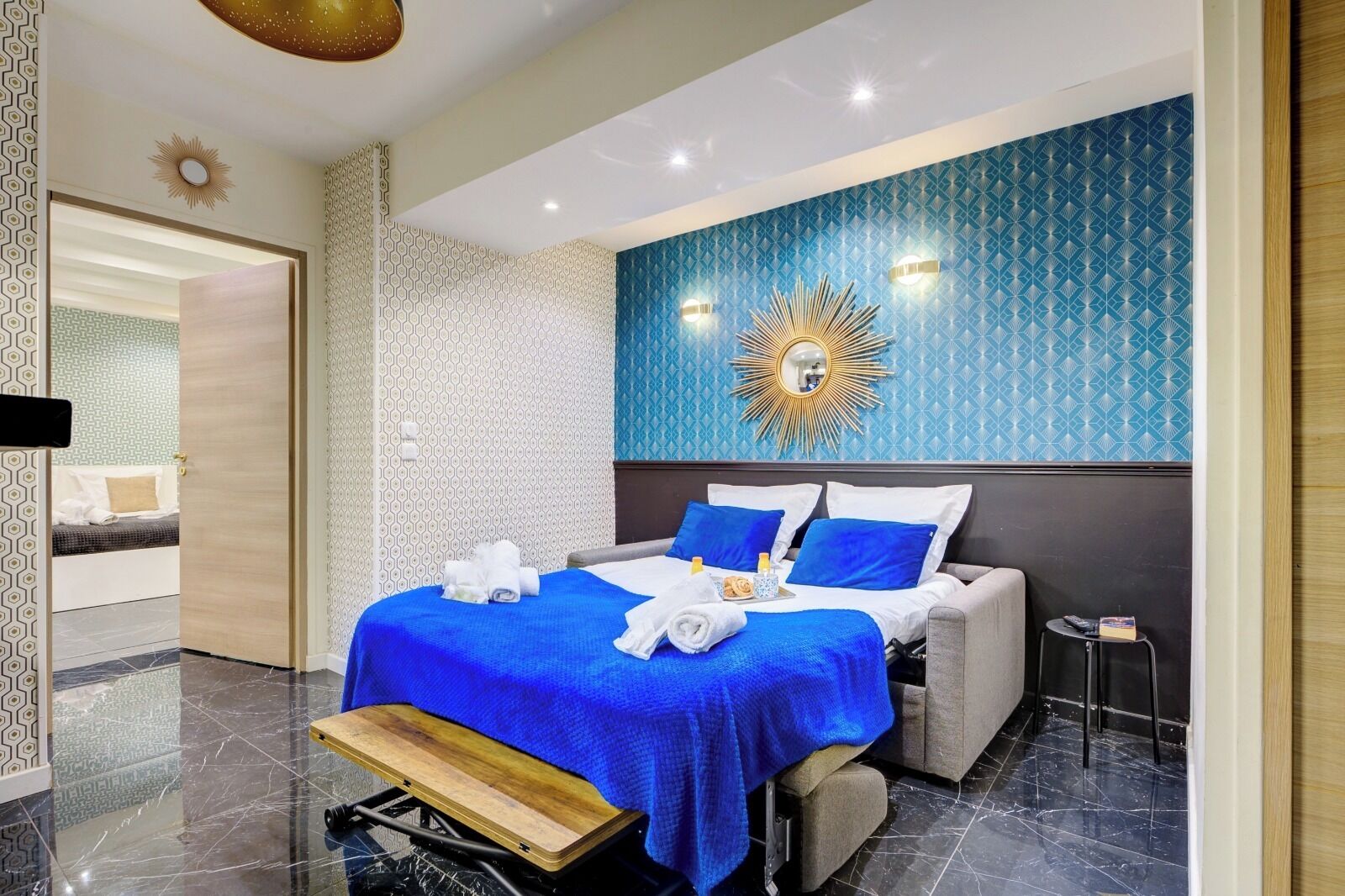 A modern hotel room with a large bed, a bench, a small table, and a decorative wall.