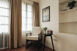 A room with a chair, a small table with a plant and a radio, a framed picture on the wall, and a window with curtains.