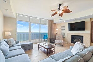 Ein geräumiges Wohnzimmer mit einem großen Sofa, einem Couchtisch und einem Kamin. An der Wand ist ein Fernseher angebracht und darüber ein Deckenventilator.