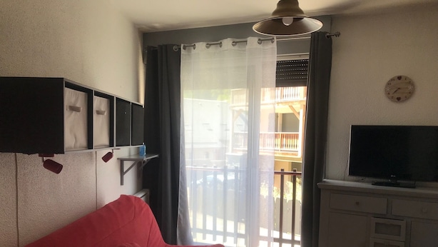 A living room with a red sofa, a television on a cabinet, a window with curtains, and a balcony visible through the window. - Studio in the mountains, 8 minutes from the slopes (Saint-Lary-Soulan)