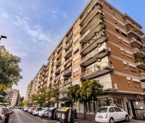 Un immeuble résidentiel à plusieurs étages, doté de balcons, avec des voitures garées et un ciel dégagé.