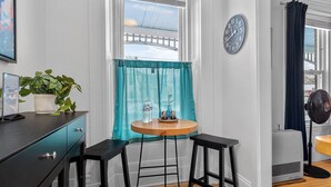 Habitación compacta con piso de madera, una pequeña mesa redonda con dos taburetes negros, un reloj de pared blanco, una planta en maceta y una ventana con cortina color turquesa.