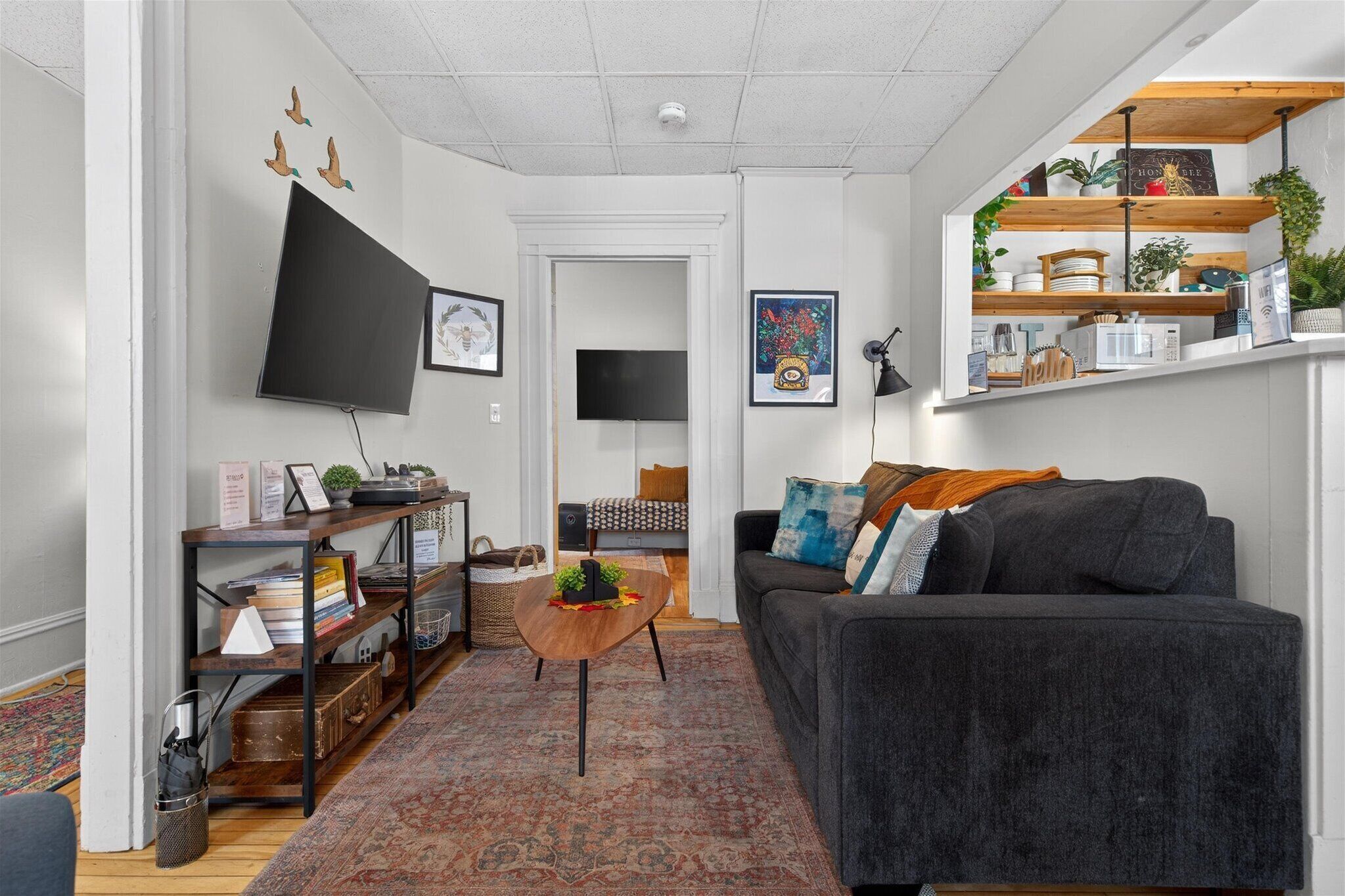 Un salon moderne avec un canapé gris foncé, une table basse en bois et un tapis à motifs. On y trouve des étagères ornées de plantes et d’objets décoratifs, ainsi qu’un téléviseur à écran plat fixé au mur.