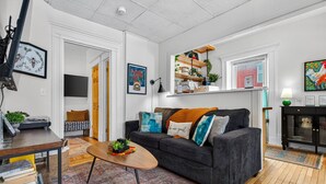 Una sala de estar moderna con un sofá gris oscuro, una mesa de centro de madera y una alfombra estampada. Se observa una estantería con objetos decorativos y un televisor de pantalla plana montado en la pared.
