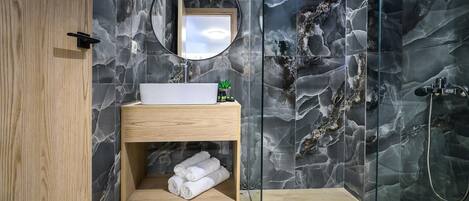 Un baño moderno con salpicadero de mármol, tocador de madera, espejo redondo y zona de ducha con mampara de vidrio.