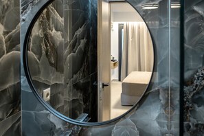 Un baño con pared de mármol, un espejo redondo y un banco beige.