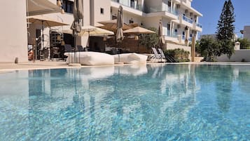 Une piscine aux eaux cristallines, bordée de chaises longues et d’ombrelles, et entourée d’un immeuble à plusieurs étages doté de balcons.