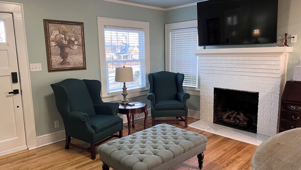 A living room with two green armchairs, a grey tufted ottoman, a white fireplace, and a mounted TV.