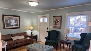 A living room with a brown leather sofa, a grey tufted ottoman, a teal armchair, and a wooden coffee table. There are two framed floral paintings on the walls, a chandelier, and a window with blinds.