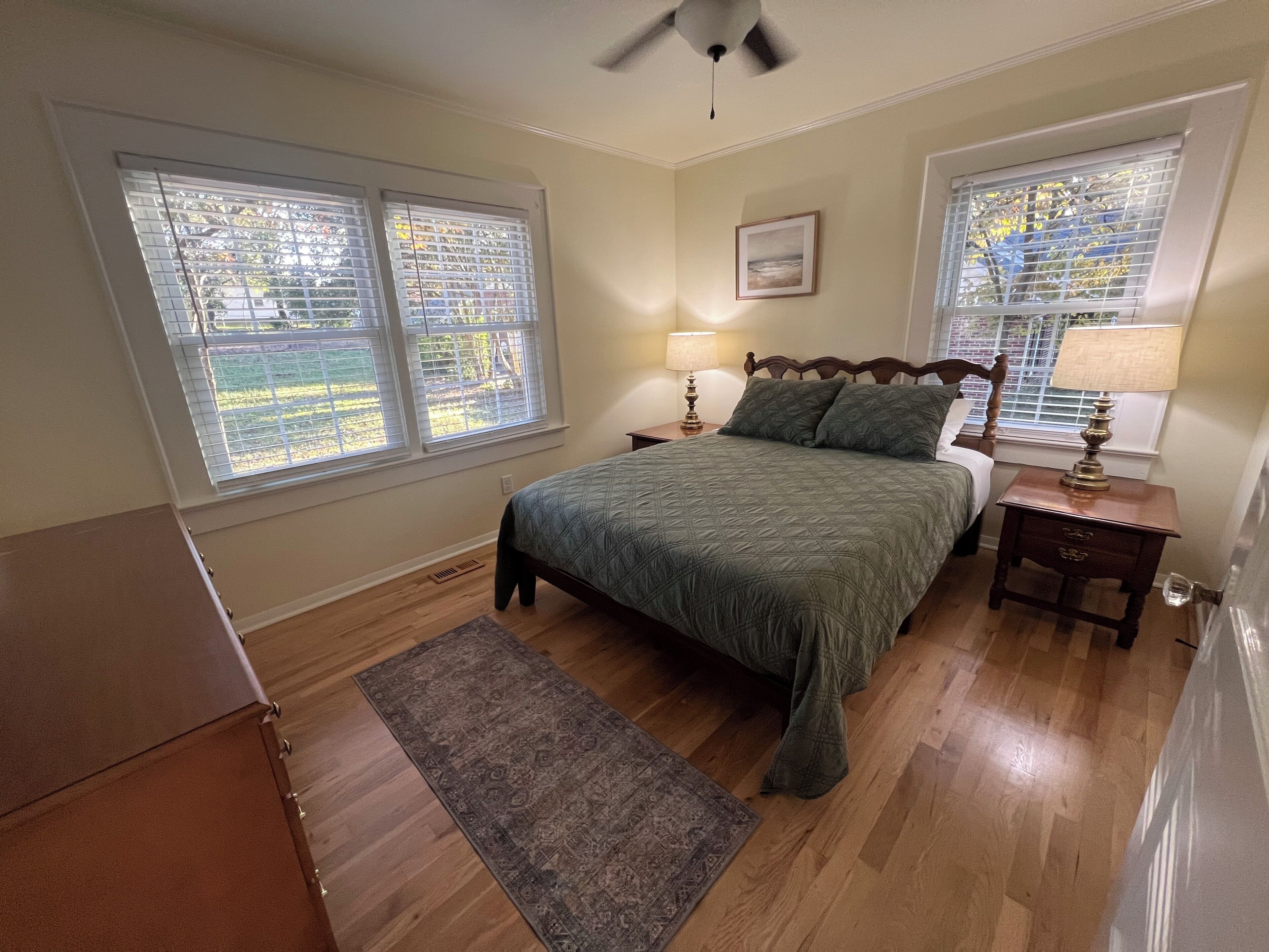A bedroom with a bed, two nightstands, a ceiling fan, and windows with blinds.