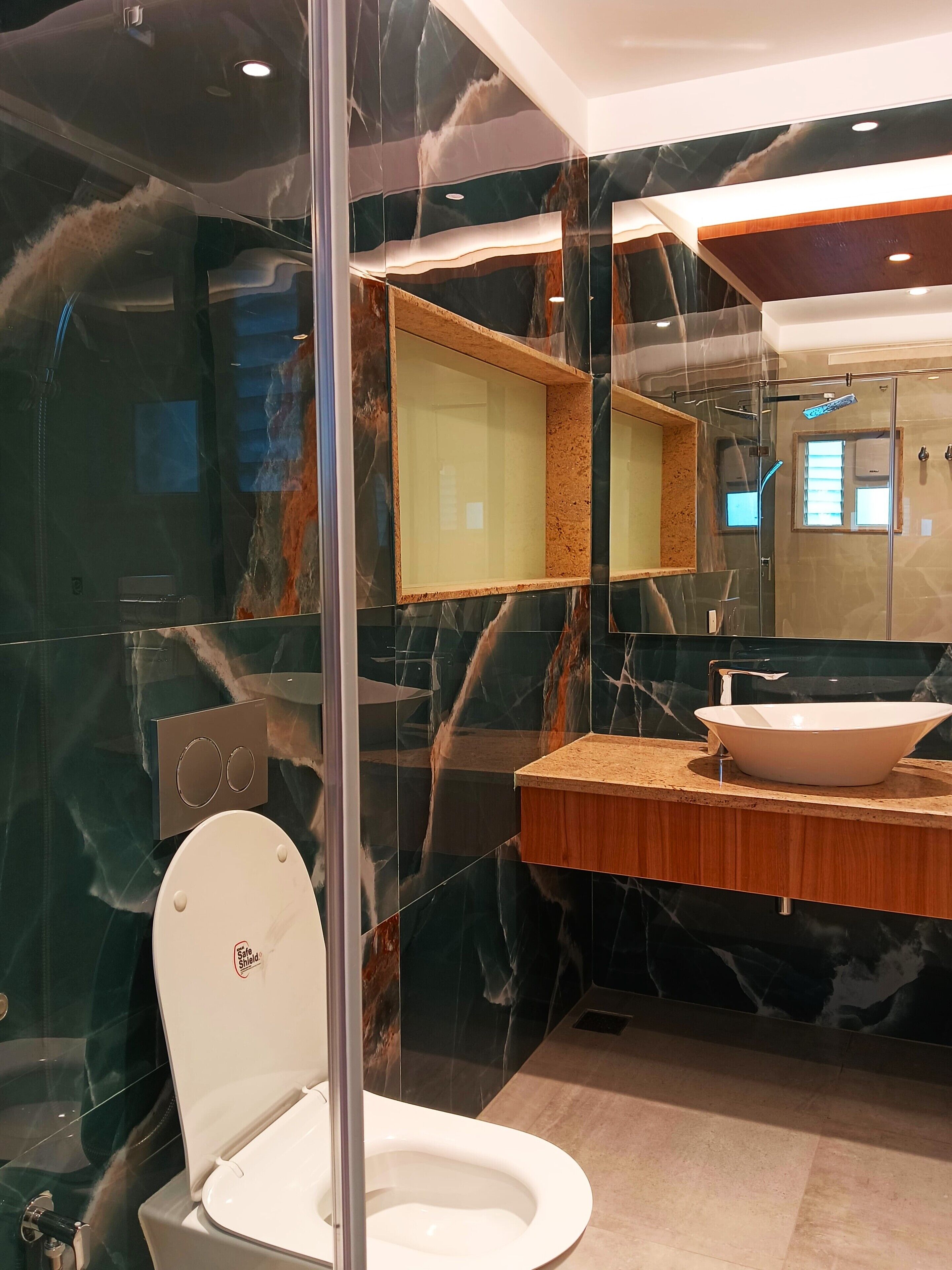 A modern bathroom with a glass-enclosed toilet, a large mirror, a wooden vanity, and a marble backsplash.