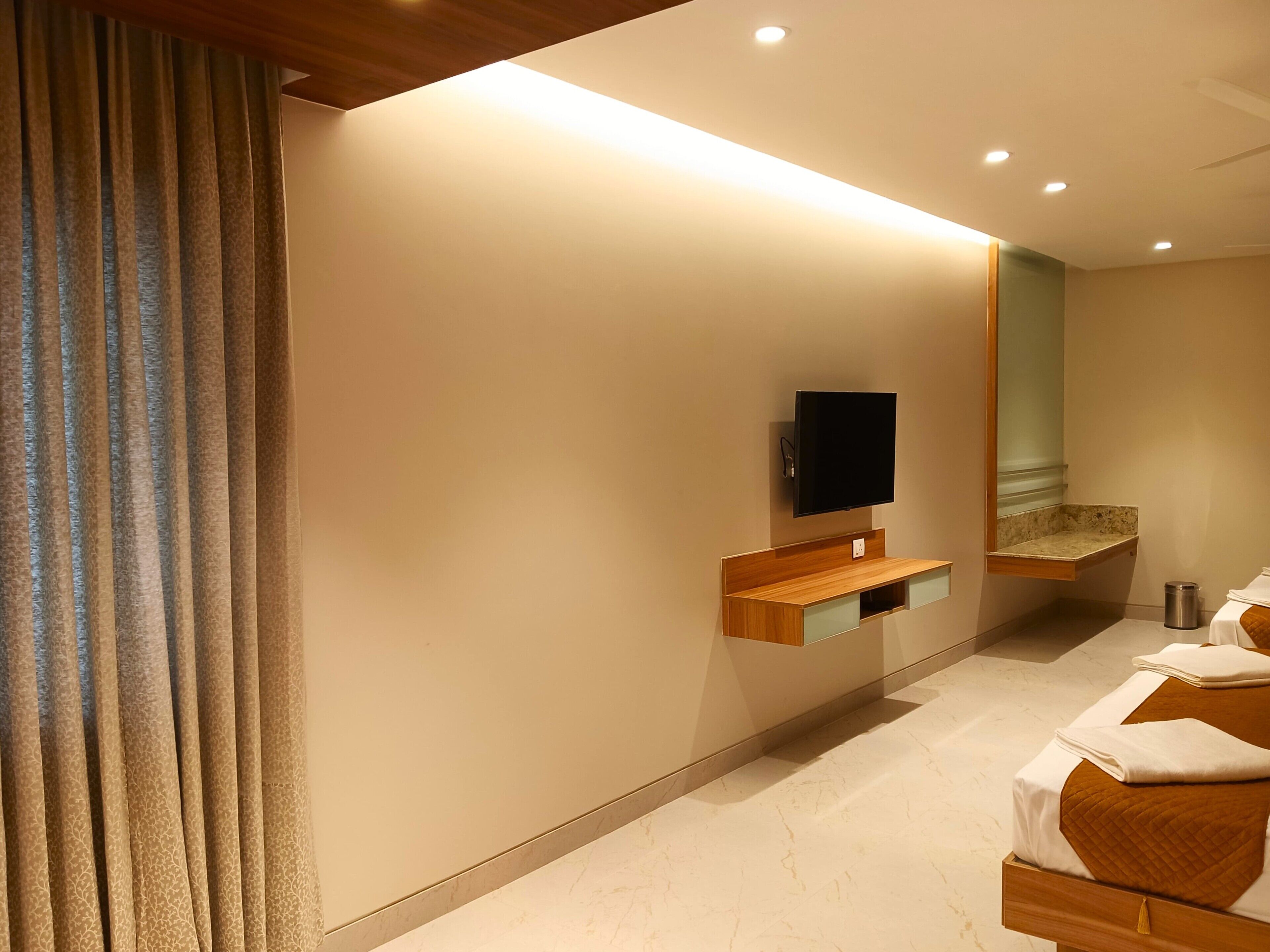Een hotelkamer met een aan de muur bevestigde televisie, een houten nachtkastje, een spiegel en een bed met een witte dekbedovertrek en bruine kussens.