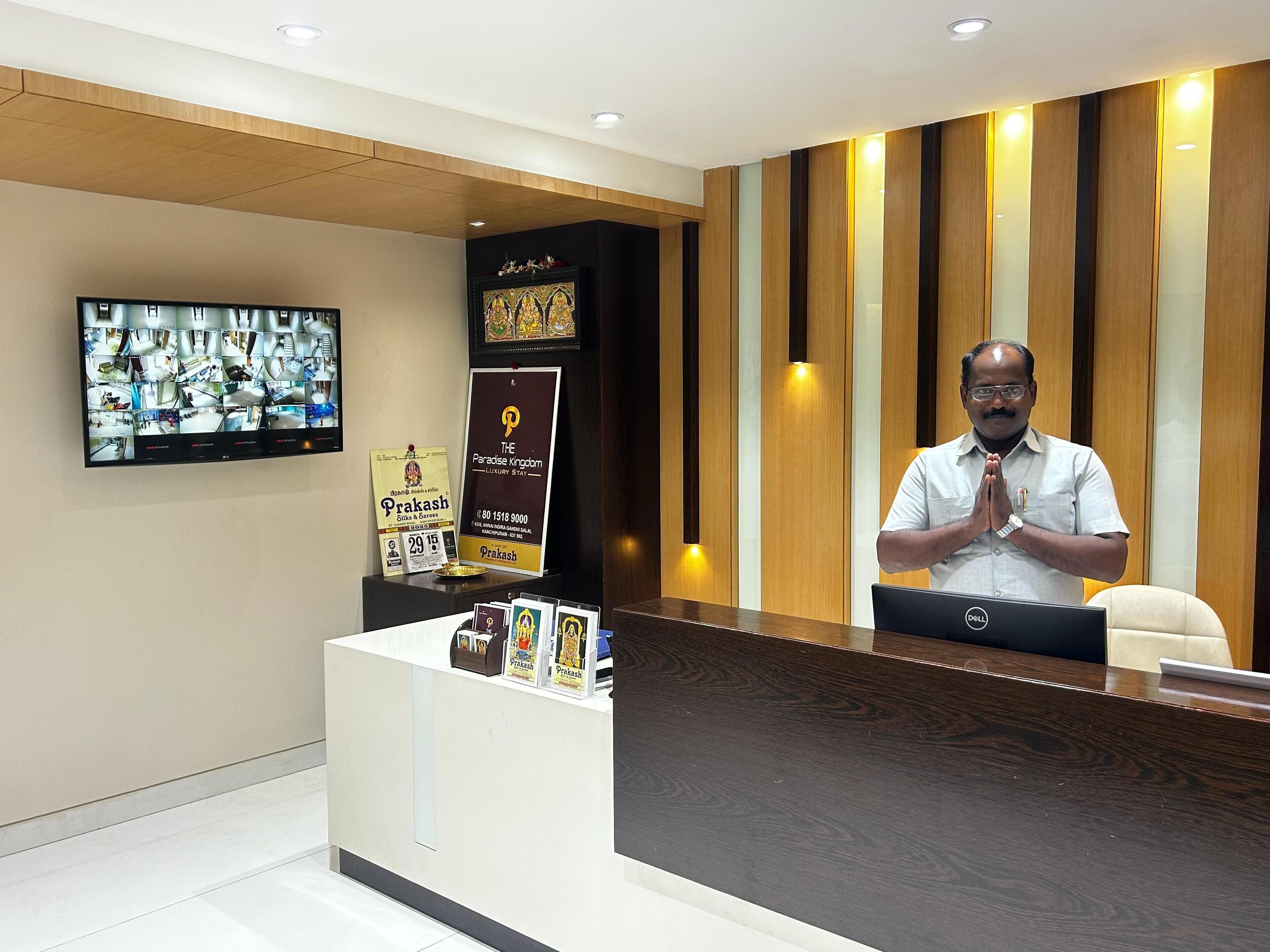 Una recepción de hotel con un monitor que muestra varias habitaciones, un cartel enmarcado y un hombre de pie detrás del mostrador.
