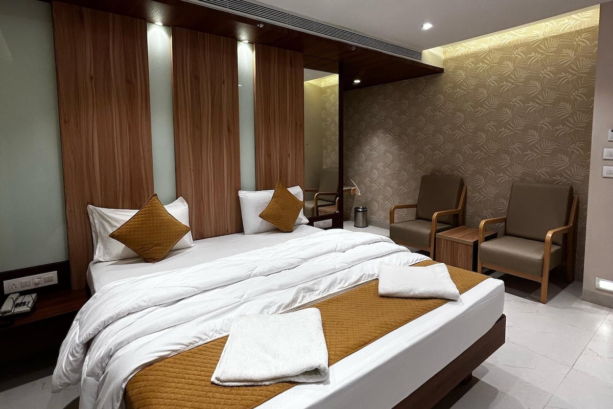 Une chambre d’hôtel moderne avec un grand lit, une tête de lit en bois et un coin salon avec des fauteuils.