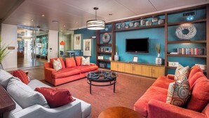 Um hall de hotel moderno com um sofá vermelho, uma poltrona cinza e uma mesa de centro. Há uma TV fixada na parede e uma estante com objetos decorativos.