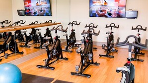 Uma academia com bicicletas ergométricas organizadas em fileiras, uma grande bola de exercícios e uma televisão exibindo conteúdo de fitness.