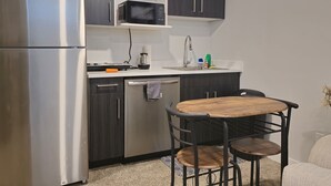 Une cuisine compacte avec des meubles de couleur sombre, un réfrigérateur en acier inoxydable, un four à micro-ondes, un évier et un petit coin repas avec une table en bois et des chaises en métal.