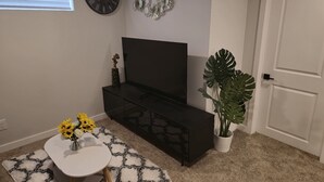 Un salon moderne avec un meuble de rangement noir pour les appareils électroniques, une table ronde blanche ornée de fleurs jaunes, une grande plante en pot et des œuvres d’art décoratives accrochées au mur.