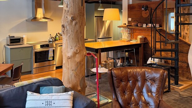 A loft-style living space with a high ceiling, exposed wooden beams, a central wooden column, a dining area with a table and chairs, a kitchen area with modern appliances, and a staircase leading to an upper level. - Cozy In Town Manchester Studio w/ Loft Bedroom. (Manchester)