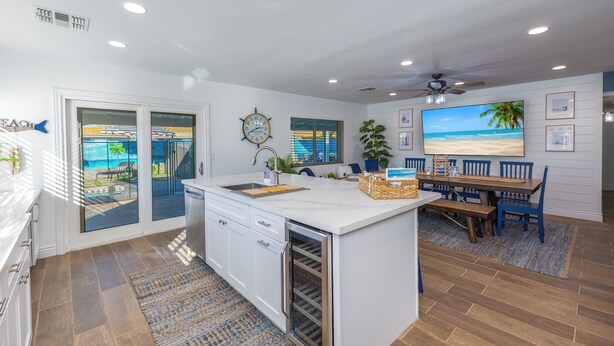 A modern kitchen with a large island, a dining area with blue chairs, a ceiling fan, and a view of the beach through the glass doors. - Life can be a Beach (Gilbert)