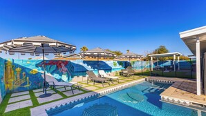 A swimming pool with lounge chairs and umbrellas, surrounded by a mural and palm trees. - Life can be a Beach (Gilbert)
