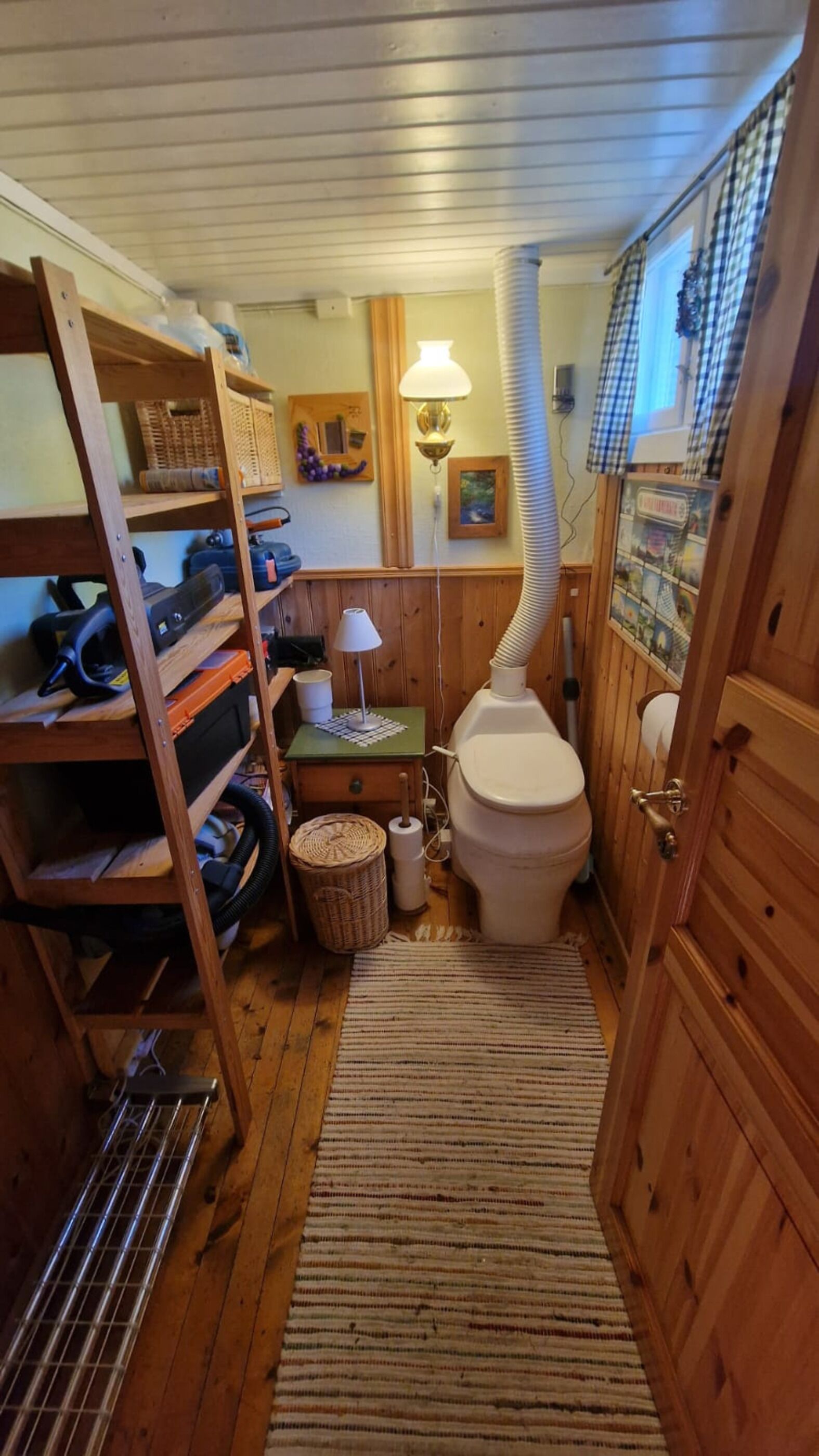 A compact bathroom with a wooden floor, a toilet, a sink with a mirror, a shelf with tools, and a lamp.