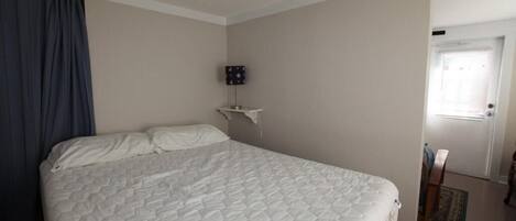 A bedroom with a large bed, a nightstand, a wall-mounted lamp, and a door leading to another room.