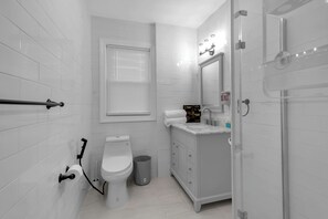 A modern bathroom with white tiles, a toilet, a sink with a mirror, and a shower area. - Dream House 3-bedroom house with AC and WiFi Near Niagara Falls (Niagara Falls)