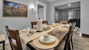 A dining area with a long table set for six, a painting on the wall, and a modern kitchen in the background.