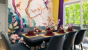 Un comedor con un mural de aves y flores, una mesa dispuesta con platos rojos y dorados, y vistas al verdor exterior.