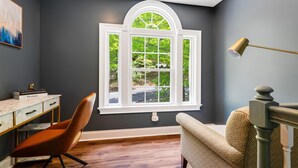 Una habitación con un amplio ventanal de arco, un escritorio con cajones, una silla y un sillón estampado.