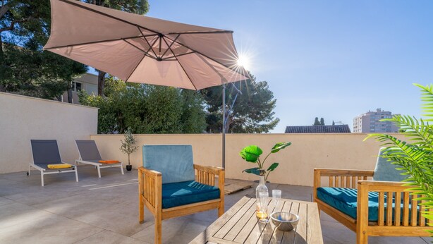 Una terraza en la azotea con muebles de madera, una mesa con un jarrón y vasos, y un gran parasol que da sombra.