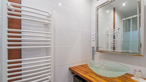 Un baño con lavamanos de vidrio, encimera de madera, azulejos blancos, cabina de ducha con mampara de vidrio y un perchero blanco.