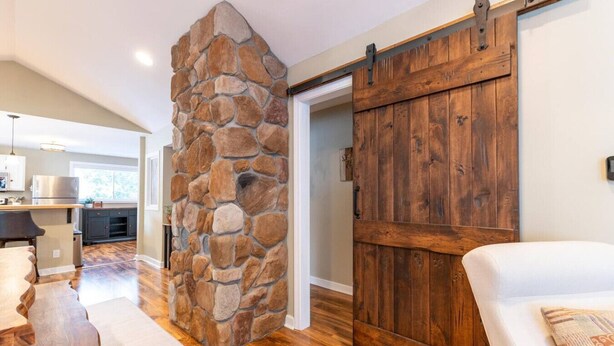 A room with a stone wall, wooden flooring, a sliding barn door, and a white sofa. - FAMILY-FRIENDLY, King Bed, Outdoor Space, Pet-Welcome - Intervale Lodge (Intervale)
