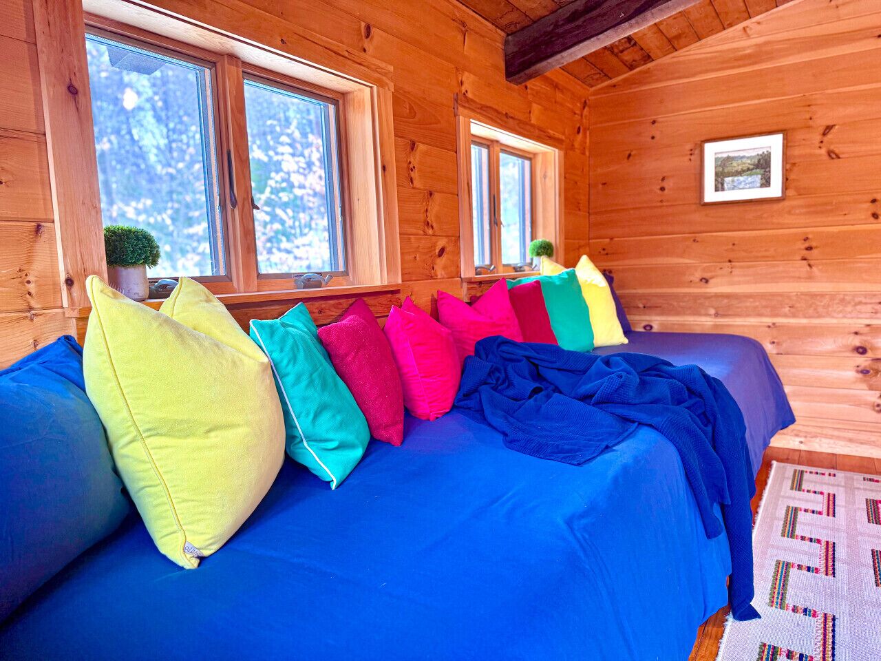 A wooden cabin room with a bed, colorful pillows, and two windows.